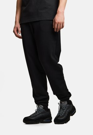 Osoba ubrana w czarne spodnie dresowe i czarno-szare sportowe buty stojąca na jasnym tle.