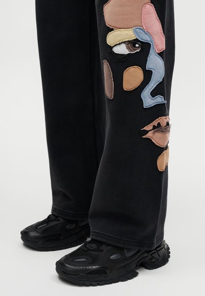 Μαύρο παντελόνι από ύφασμα με πολύχρωμα κεντημένα μπαλώματα σε διάφορα σχήματα. Φορεμένο με μαύρα παπούτσια με υφή και χοντρή σόλα.