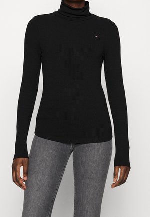 Personne portant un pull noir ajusté à col roulé côtelé et un jean gris, debout devant un fond blanc uni.