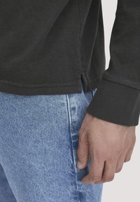 Dunkelgraues Langarmshirt mit seitlichen Schlitzen, kombiniert mit hellblauen Jeans. Nahaufnahme einer Hand, die auf den Jeans ruht.