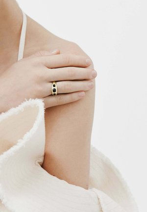 Anillo de oro con dos piedras negras, llevado en una mano que descansa sobre un hombro. En el fondo se asoma un tejido suave y texturizado.