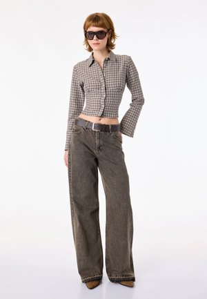 Donna con occhiali da sole neri oversize, camicia a quadri corta con bottoni, jeans sbiaditi a gamba larga, cintura nera e scarpe marroni appuntite su sfondo bianco.