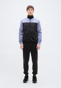 Αθλητική ζακέτα σε μαύρο και λιλά, με ψηλό γιακά και κλείσιμο με φερμουάρ. Ομαλό ύφασμα, συνδυασμένο με μαύρο παντελόνι και αθλητικά παπούτσια σε αντίστοιχο χρώμα.