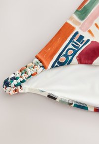 Tissu coloré à motif avec un détail de sangle tressée, probablement partie d’un maillot de bain ou d’un bas de bikini.