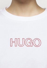 Biała koszulka z okrągłym dekoltem, z czerwoną ramką wokół napisu „HUGO” na środku klatki piersiowej, noszona przez osobę z widoczną szyją i obojczykiem.