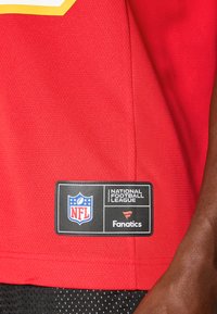 Sarkans NFL krekls ar teksturētu audumu, kurā ir dzeltena un balta dizaina akcenti. Ietver etiķeti ar NFL un Fanatics zīmola apzīmējumu.
