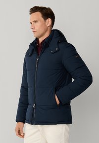 Hombre con chaqueta acolchada azul marino con capucha, de pie con una mano en el bolsillo contra un fondo liso.