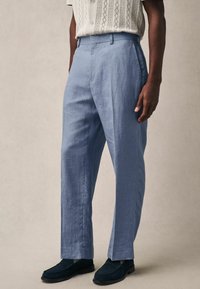 Pantalones de lino azul claro con un corte relajado, que presentan pliegues delanteros y bolsillos laterales, combinados con mocasines oscuros.