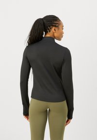 Tricou sportiv negru cu mânecă lungă, guler cu fermoar, design ajustat și textură netedă. Asortat cu colanți de culoare verde măsliniu.