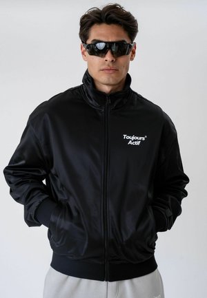 Uomo che indossa una giacca nera con zip con la scritta "Toujours Actif" sul petto e occhiali da sole scuri avvolgenti, in piedi con le mani nelle tasche.