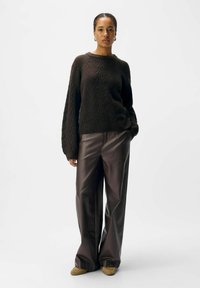 Dunkelbrauner gestrickter Pullover mit strukturiertem Muster, kombiniert mit lockeren braunen Lederhosen und hellbraunen Schuhen. Das Model steht vor einem weißen Hintergrund.