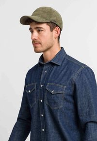 Denim shirt in dark blue with a pointed collar, two chest pockets, and button closure. Olive green corduroy cap with a curved brim.