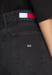 Černé džíny z denimu s zadní kapsou a barevným „TOMMY JEANS“ štítkem na pasu, který obsahuje červené, bílé a zelené akcenty.