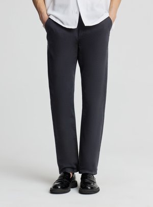 Persona che indossa pantaloni neri a gamba dritta, mocassini in pelle nera e una camicia bianca con le mani nelle tasche su uno sfondo uniforme.