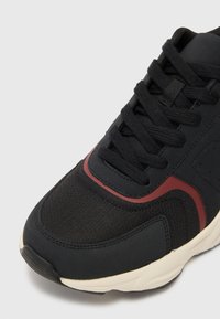 Black sneaker with a textured upper, featuring a red accent and gray midsole. Laces are black, and the sole shows a slight curve.