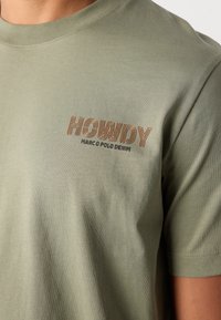 Olíva zöld pamut póló, melynek bal mellkasán narancssárga és fekete betűkkel szerepel a "HOWDY" felirat, sima textúrájú és rövid ujjú.