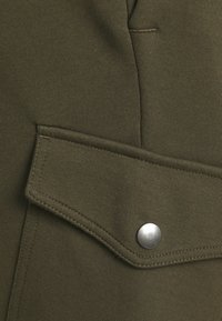 Olivově zelená látka, která obsahuje texturovanou kapsu s kovovým zapínáním a detailem šití, z kusu oděvu.