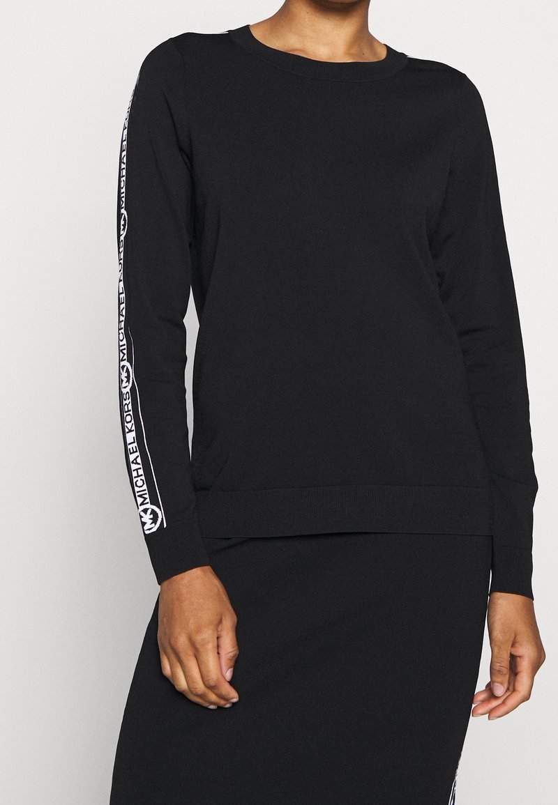 Personne portant un haut noir à manches longues et une jupe avec une bande blanche portant le logo "Michael Kors" le long des manches, sur un fond uni.