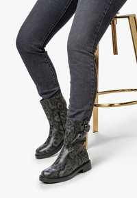 Botas de tobillo con estampado de serpiente negra, con punta redonda y detalle decorativo de hebilla, combinadas con jeans oscuros ajustados y un taburete dorado.