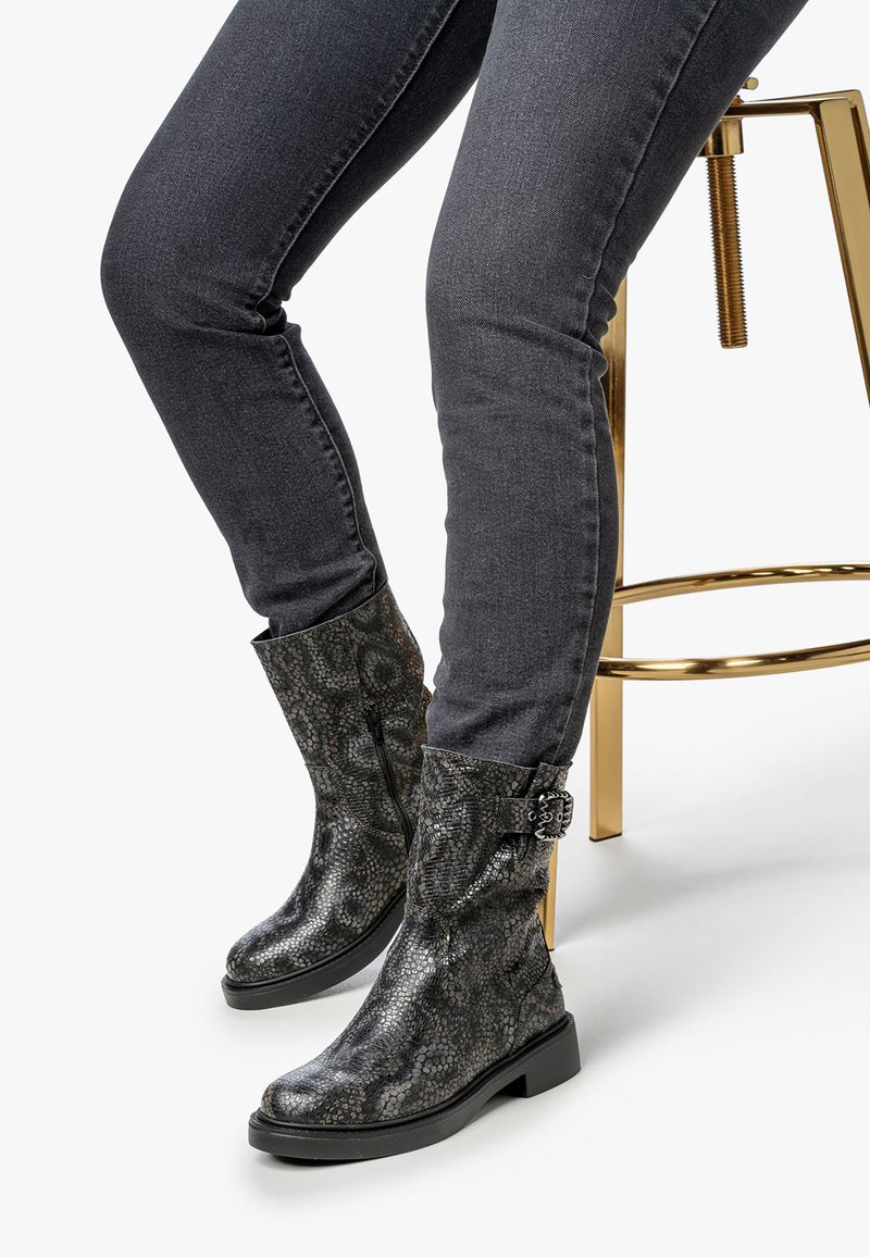 Botas de tobillo con estampado de serpiente negra, con punta redonda y detalle decorativo de hebilla, combinadas con jeans oscuros ajustados y un taburete dorado.
