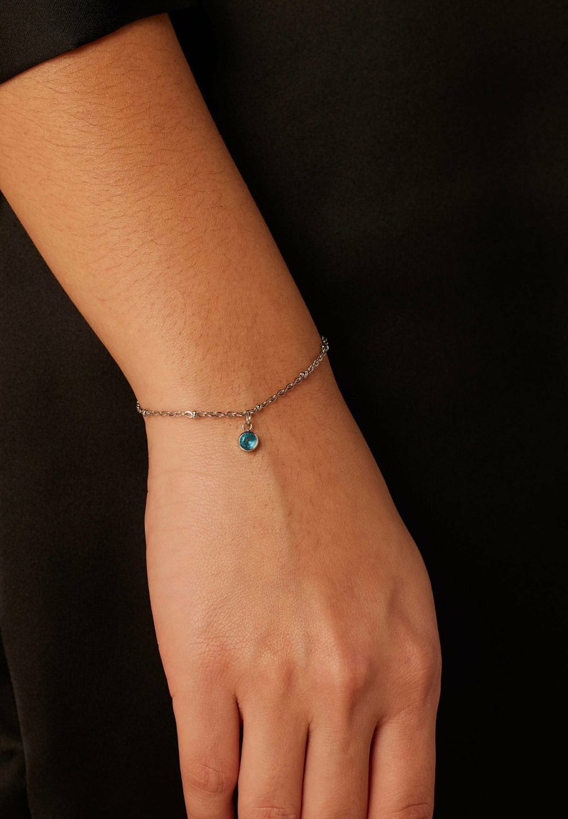 Zilveren kettingarmband met een ronde, lichtblauwe edelsteenhanger, gedragen om een pols met textuur, tegen een donkere achtergrond.