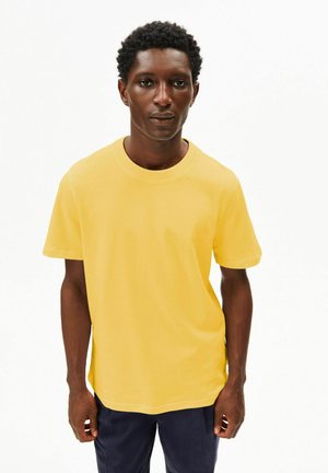 Giovane uomo con capelli ricci corti che indossa una maglietta gialla semplice e pantaloni scuri, in piedi contro uno sfondo bianco.