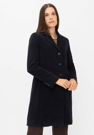 Frau mit glattem braunem Haar, die einen schwarzen, geknöpften Mantel trägt und mit verschränkten Händen vor einem schlichten weißen Hintergrund steht.