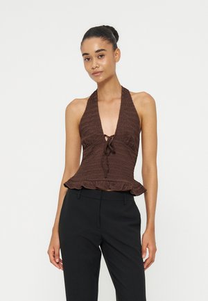 Jeune femme aux cheveux foncés portant un haut marron sans manches avec un nœud à l'avant et un pantalon noir, debout devant un fond blanc uni.