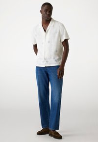 Short-sleeve white linen shirt featuring light blue patterns, paired with blue denim jeans and brown moccasin shoes. Standing pose.
