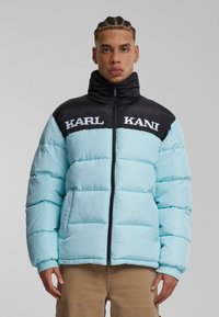 Lyseblå pufferjakke med sort krave, forsynet med hvid "KARL KANI" logo, quiltet design og elastiske manchetter. Lynlåslukning foran.