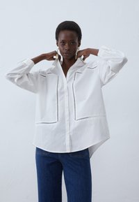 Witte overhemd met lange mouwen, voorzien van decoratieve zwarte stiksels aan de voorkant. Gecombineerd met blauwe spijkerbroek.