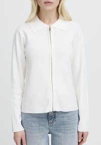 Femme portant un pull à fermeture éclair blanc à manches longues avec un col et un jean bleu clair, sur un fond blanc uni.