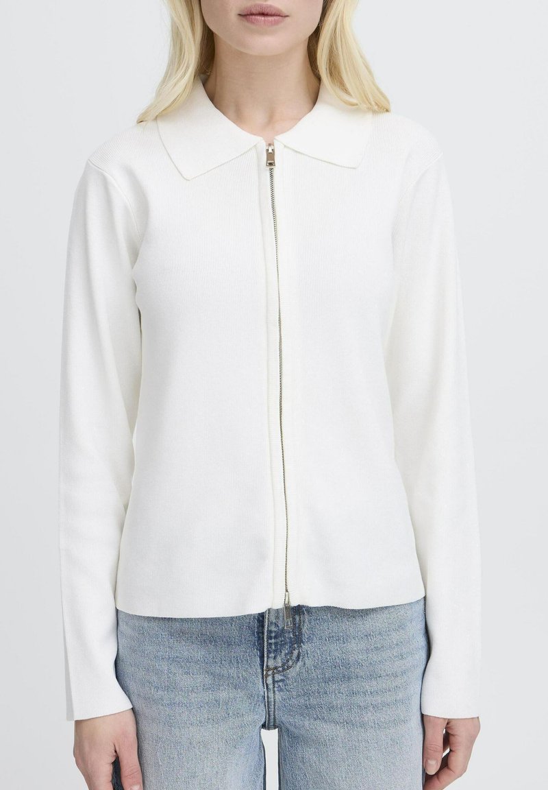 Femme portant un pull à fermeture éclair blanc à manches longues avec un col et un jean bleu clair, sur un fond blanc uni.