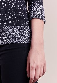 Top nero con un motivo floreale testurizzato e dettagli bianchi, contrastato da un bordo inferiore nero uniforme. Maniche lunghe con polsini arrotolati.