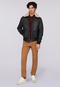 Černá kožená bomber bunda s béžovým ovčím límcem, dvěma předními kapsami, žebrovanými manžetami, v kombinaci s hnědými kalhotami a bílými teniskami.