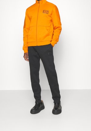 Homme portant une veste zippée orange vif avec le logo EA7 et un pantalon de survêtement noir avec une poche à patch, debout devant un fond blanc.