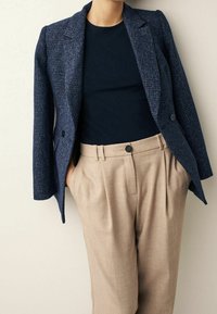 Persona con una chaqueta azul marino texturizada sobre una camiseta negra y pantalones beige de talle alto con pliegues, con las manos en los bolsillos, de pie contra una pared clara.