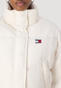 Giacca a vento color crema con collo alto, tasche laterali e una patch con logo in blu, rosso e bianco sul lato sinistro del petto.