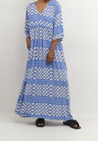 Maxiklänning i blått bomull med beige mönster, med V-hals, korta ärmar och indragen midja. Enfärgade sandaler fulländar looken.