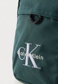 Grønn ryggsekk i stoff med teksturert overflate, toppklaff med en svart løkke, og brodert "Calvin Klein"-logo i hvitt og lys grått.