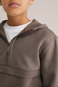 Ruskea vetoketjulla varustettu huppari, jossa on teksturoitu alaosa ja logotyyppikoristeet. Hupullinen ja sileäpintainen kangas. Päällä valkoinen paita.