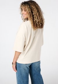Beige oversized top met korte mouwen, zachte stof, gebogen zoom, gecombineerd met lichtblauwe wijde jeans. Geen zichtbare patronen of logo's.