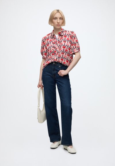 Femme aux cheveux blonds et courts, portant une chemise rouge à motifs et manches courtes, un jean bleu foncé, des chaussures blanches, tenant un sac à main blanc, se tenant devant un fond blanc.