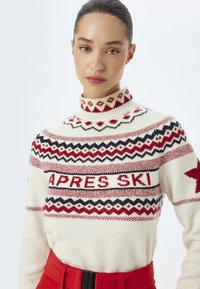 Stickad tröja i krämfärgad med röda och svarta geometriska mönster. Har en hög krage och texten 'APRES SKI' överst på framsidan.
