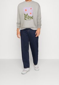 Persona che indossa una felpa grigia con una grafica di fiore rosa e jeans blu scuro, in piedi su uno sfondo bianco con sneaker bianche.