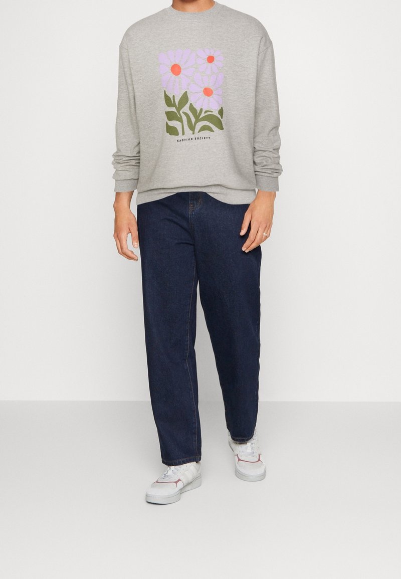 Persona che indossa una felpa grigia con una grafica di fiore rosa e jeans blu scuro, in piedi su uno sfondo bianco con sneaker bianche.