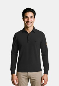Zwarte polo met lange mouwen, drie knopen en een klein logo op de borst. Zacht materiaal en geribbelde manchetten. Draagt met lichtgekleurde broeken.