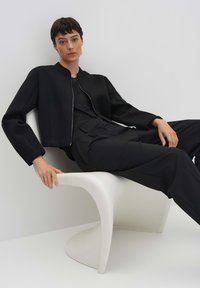 Veste noire courte avec une texture lisse et une fermeture éclair, assortie à un pantalon noir, portée par une personne assise sur une chaise sculpturale blanche.