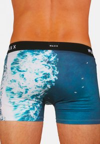 Boxers en tissu imprimé vague, principalement turquoise et blanc, avec une ceinture élastique noire portant l'étiquette "WAXX."