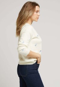 Pull crème avec broderies florales bleues, texture douce, col rond et manches longues. Associé à un jean bleu foncé.